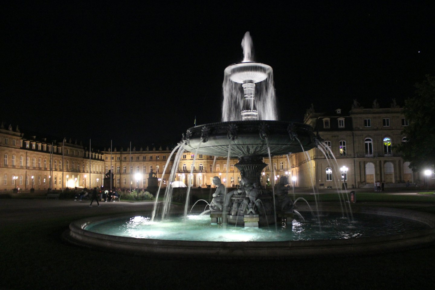 Ein mehrstufiger Steinbrunnen fließt nachts auf einem Stadtplatz.