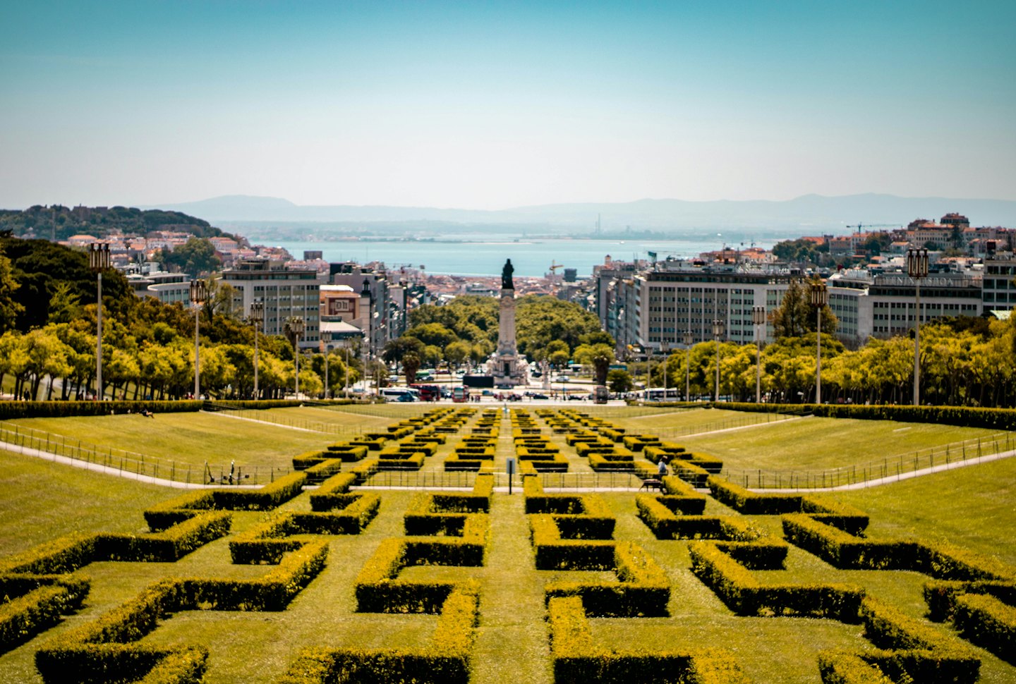 Geometrische Gartenhecken führen zu einem Denkmal mit Blick auf eine städtische Skyline.